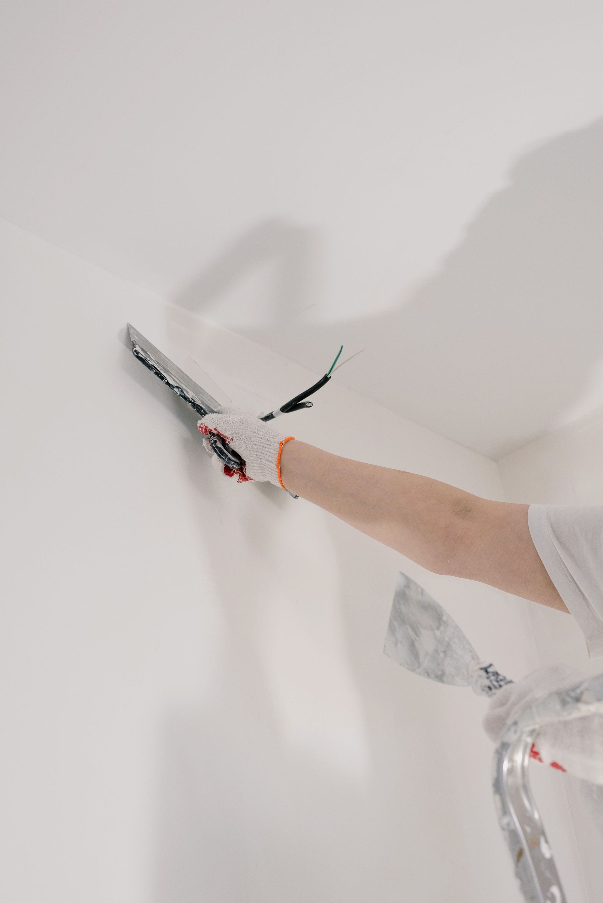 Crop unrecognizable male in gloves holding tool while standing on ladder and renovating walls in flat